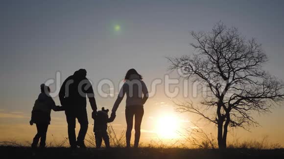 日落时幸福的家庭在一棵大树附近的草地上散步的剪影视频的预览图