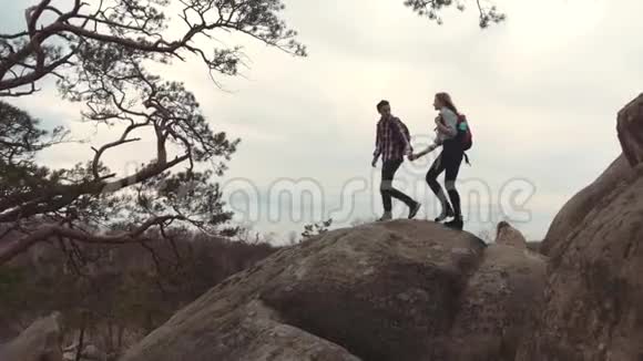 一对年轻情侣拿着背包手牵着手沿着高高的石头山爬山旅游视频的预览图