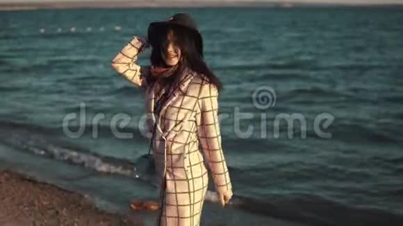 年轻女子在水边享受一个温暖的秋天下午穿着秋装戴着帽子的女孩在日落时沿着海岸散步视频的预览图