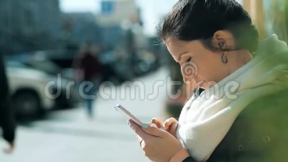 美女时尚年轻女性在街上坐在长凳上用智能手机发短信视频的预览图