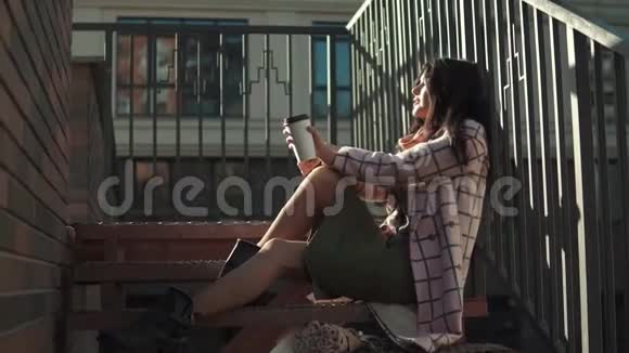 希普斯特女孩在现代建筑背景下的台阶上喝咖啡秋衣少女视频的预览图