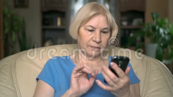 开朗好看的高级女性坐在家里的椅子上利用她的智能手机浏览视频的预览图