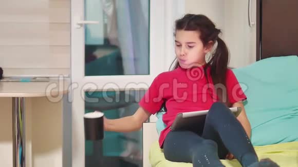 小女孩在玩数字平板电脑吃三明治小女孩坐在床上玩数字平板电脑有零食视频的预览图