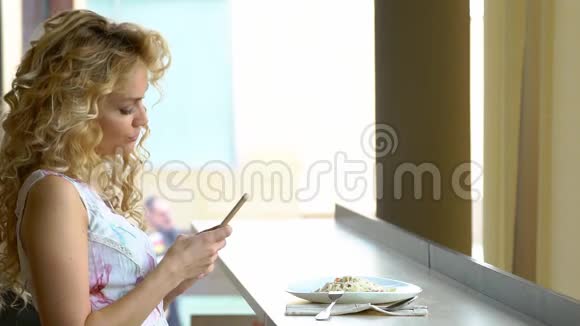 午餐休息时一个年轻的金发女孩坐在咖啡馆里用智能手机数码相机为自己画像视频的预览图