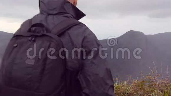 年轻的男徒步旅行者穿着雨衣背包伸向山顶举起双手男子游客站在视频的预览图