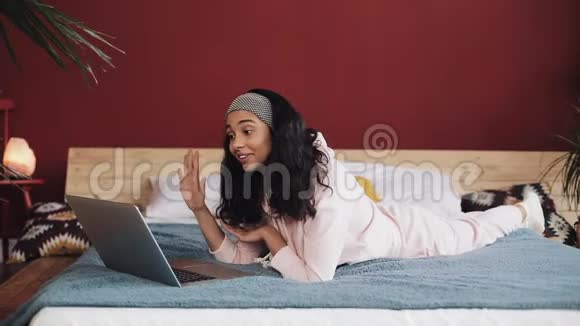 快乐的非裔美国女孩躺在靠窗的床上和她的朋友在视频电话中愉快地聊天视频的预览图