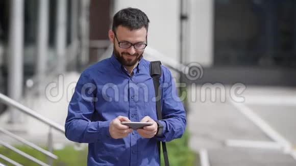 穿着蓝色衬衫和棕色裤子的英俊大胡子男子站在户外用他的手机输入信息视频的预览图