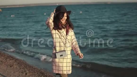 年轻女子在水边享受一个温暖的秋天下午穿着秋装戴着帽子的女孩在海滨散步视频的预览图