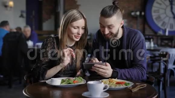 快乐的一对夫妇一起吃午饭看智能手机男人和女人用数码小玩意笑照片视频的预览图