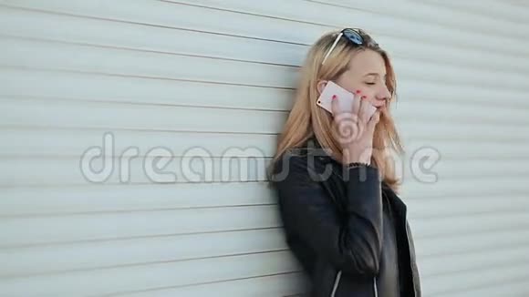漂亮的金发女孩穿着皮夹克和太阳镜在户外用手机说话在一个背景下视频的预览图