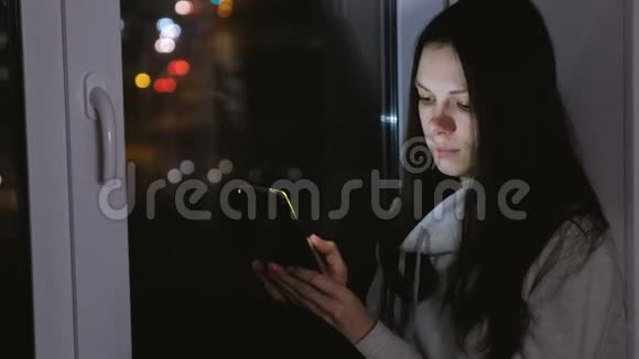 女人在黑暗的夜晚坐在窗台上用手机浏览网页视频的预览图