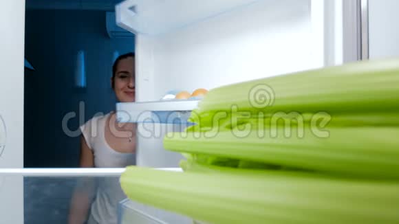 4k段快乐微笑的女人在冰箱里寻找食物和咬芹菜视频的预览图