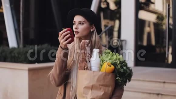 年轻漂亮的女人穿着时尚的外套站在街上拿着一包产品她拿着红苹果视频的预览图