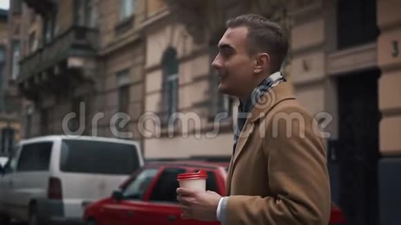 一个穿斗篷的年轻商人在多云的天气里走在街上喝着咖啡慢动作视频的预览图