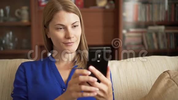 漂亮的年轻女士穿着蓝色衬衫坐在客厅的沙发上用智能手机自拍视频的预览图
