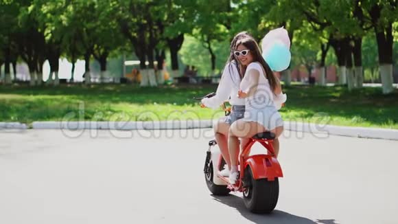 夏天两个穿牛仔短裤的漂亮女孩在公园里骑着电动滑板车视频的预览图