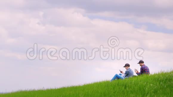 两个十几岁的男孩坐在一片绿色的草地上用手机在蓝天的背景下视频的预览图