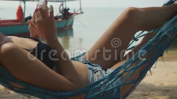 年轻女子慢动作躺在沙滩上用智能手机吊床上视频的预览图