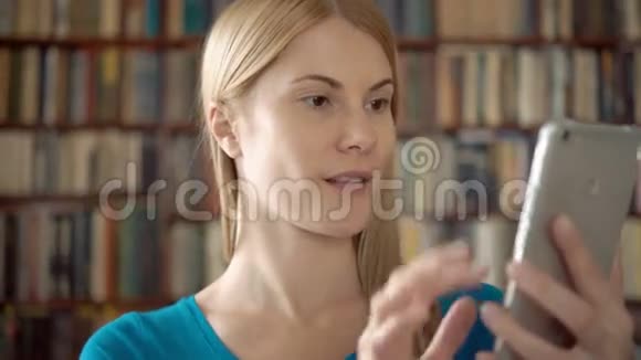 快乐的微笑学生站在图书馆用她的智能手机通过Skype与朋友聊天视频的预览图