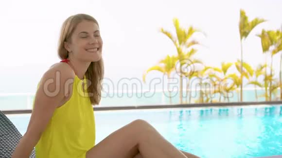 一位穿着黄色衣服坐在绿松石水游泳池旁的日光浴池里的美丽梦幻的年轻女子视频的预览图