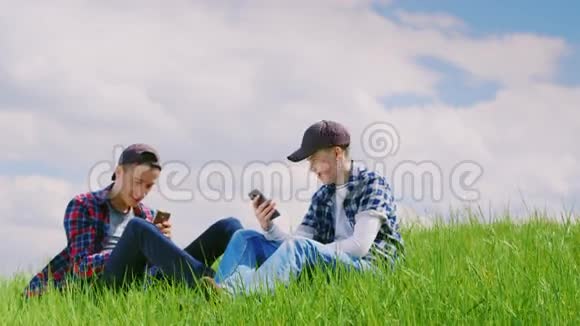 两个十几岁的男孩在大自然中交流他们坐在绿色的草地上用电话在一个视频的预览图