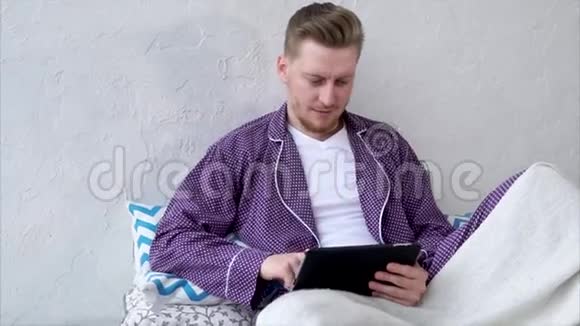 一个穿着睡衣的年轻人通过互联网在游戏中玩便携式平板电脑视频的预览图