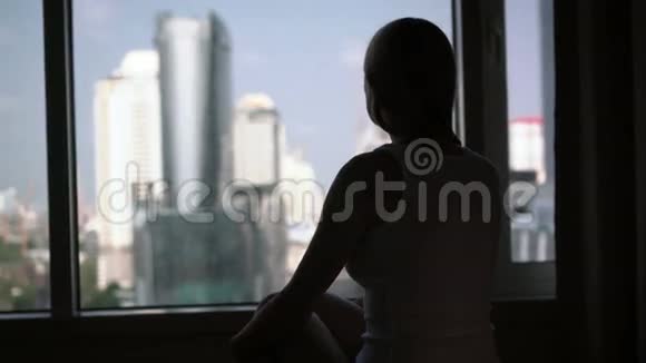 年轻女子坐在床上望着窗外的剪影城外摩天大楼景观视频的预览图