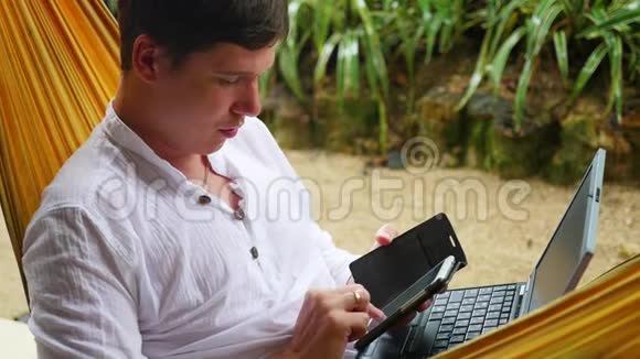 在笔记本电脑和智能手机上工作的年轻人躺在吊床上视频的预览图