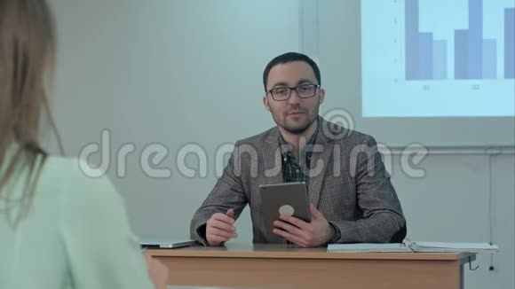 男老师拿着数码平板电脑坐在学生面前对着摄像机说话视频的预览图