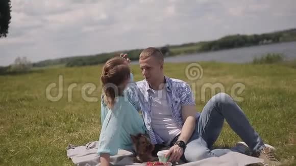 在河边的草地上约会恋爱的年轻夫妇一个男人和一个女孩和一只狗散步视频的预览图