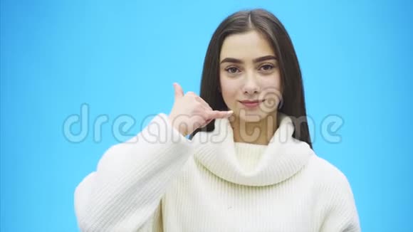 蓝色背景上美丽的年轻女孩女孩用手势显示一个电话穿着白色温暖的毛衣视频的预览图