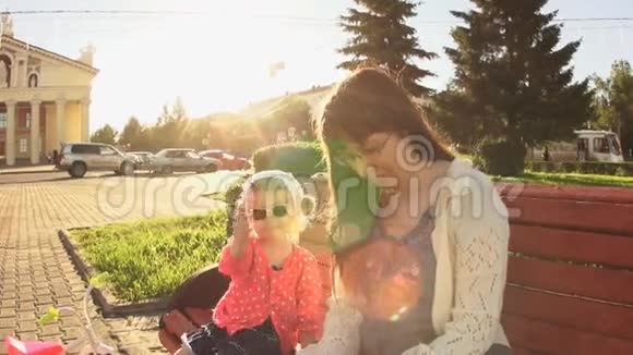 日落时分母亲和女儿正坐在靠近马路的广场上的长凳上视频的预览图