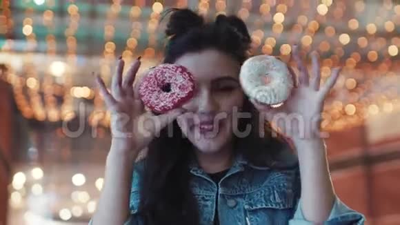 近距离观看漂亮的年轻女人用甜甜圈遮住眼睛背景上明亮的灯光玩的心情视频的预览图