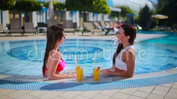 两个漂亮的黑发女郎在游泳池里喝果汁放松视频的预览图
