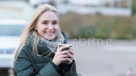 秋天微笑的年轻女子端着茶或咖啡在户外喝酒看着街上的镜头视频的预览图