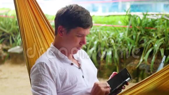 在笔记本电脑和智能手机上工作的年轻人躺在吊床上视频的预览图
