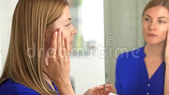 一位穿着深蓝色衬衫的美丽迷人的年轻女子在浴室的镜子里看着她的脸视频的预览图