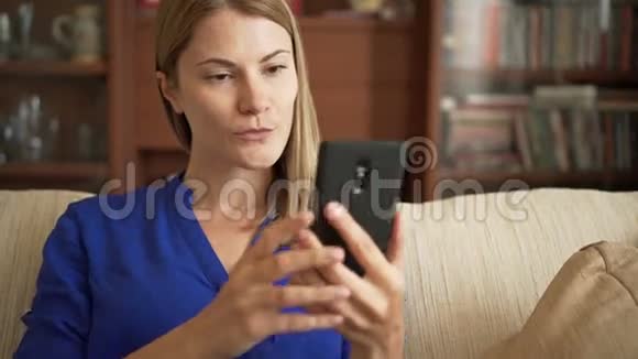 漂亮的年轻女士穿着蓝色衬衫坐在客厅的沙发上用智能手机自拍视频的预览图