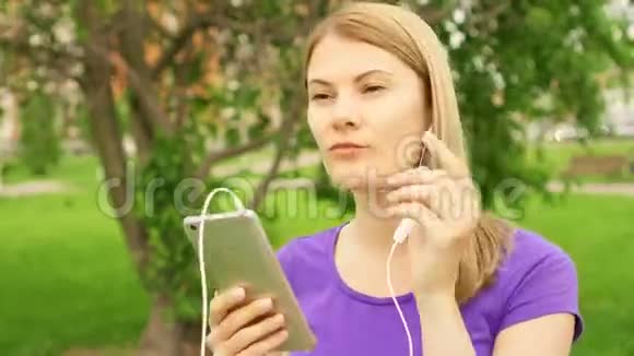 女人在公园里用耳机听手机音乐用她的智能手机跳舞唱歌视频的预览图