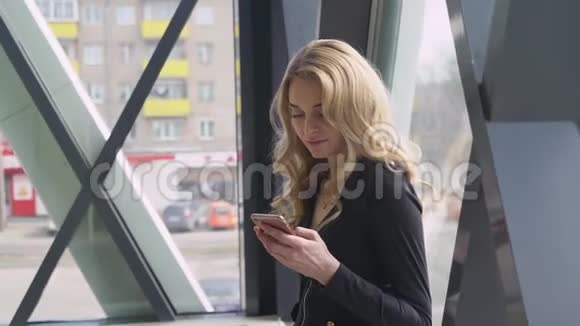 漂亮的金发女孩在明亮的窗户附近享受电话和拨号信息视频的预览图