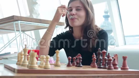 美丽的年轻女孩有着棋盘微笑着坐在咖啡馆的桌子上喝着玻璃烧杯里的茶弯着腰视频的预览图