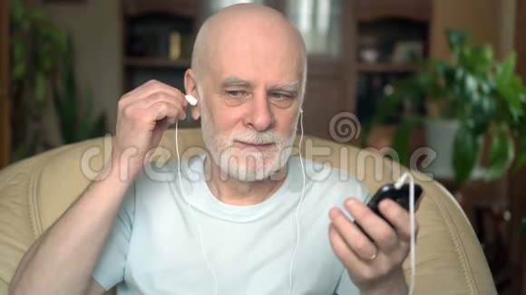 好看的老人坐在家里的椅子上智能手机带耳机听音乐视频的预览图
