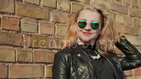 时尚的金发女郎戴着太阳镜和黑色皮夹克靠近街道上的砖墙视频的预览图