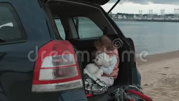 美丽的小女孩坐在河岸一辆敞篷的汽车后备箱里用智能手机视频的预览图