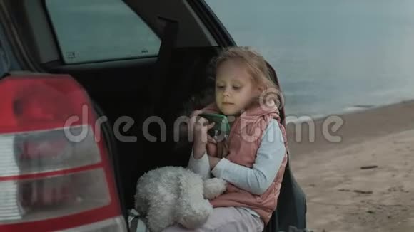 美丽的小女孩坐在河岸一辆敞篷的汽车后备箱里用智能手机视频的预览图