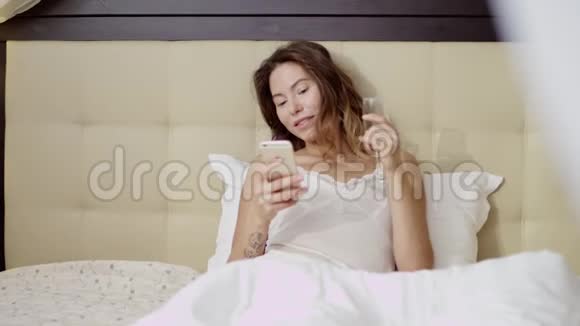 迷人的女人躺在床上拿着一杯香槟和一部智能手机视频的预览图