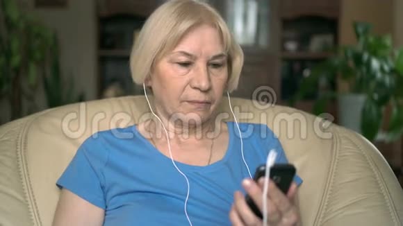 漂亮的高级女人坐在家里的椅子上智能手机带耳机听音乐视频的预览图