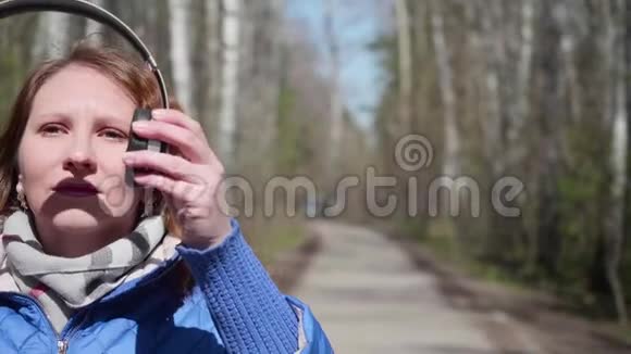 公园里的一个年轻女孩用耳机听音乐一个女人走过春天公园欣赏音乐视频的预览图