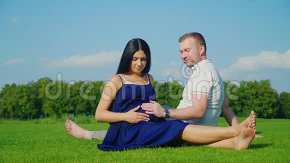 年轻的丈夫和他怀孕的妻子在公园的绿色草坪上放松视频的预览图