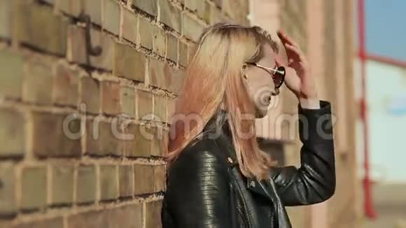 时尚的金发女郎戴着太阳镜和黑色皮夹克靠近街道上的砖墙视频的预览图